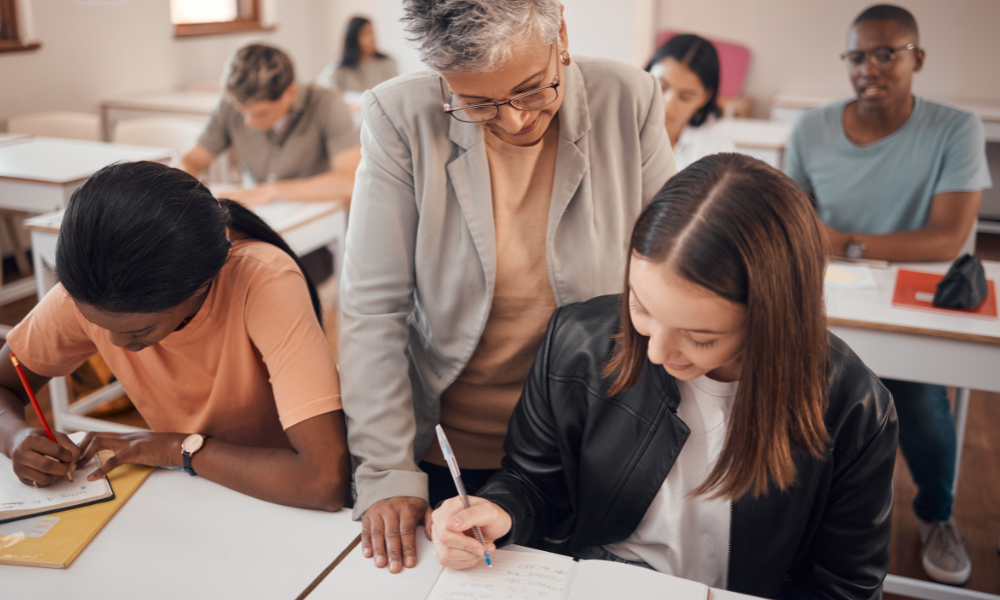Students in a classroom, focused on their work. A  teacher is guiding a female student.